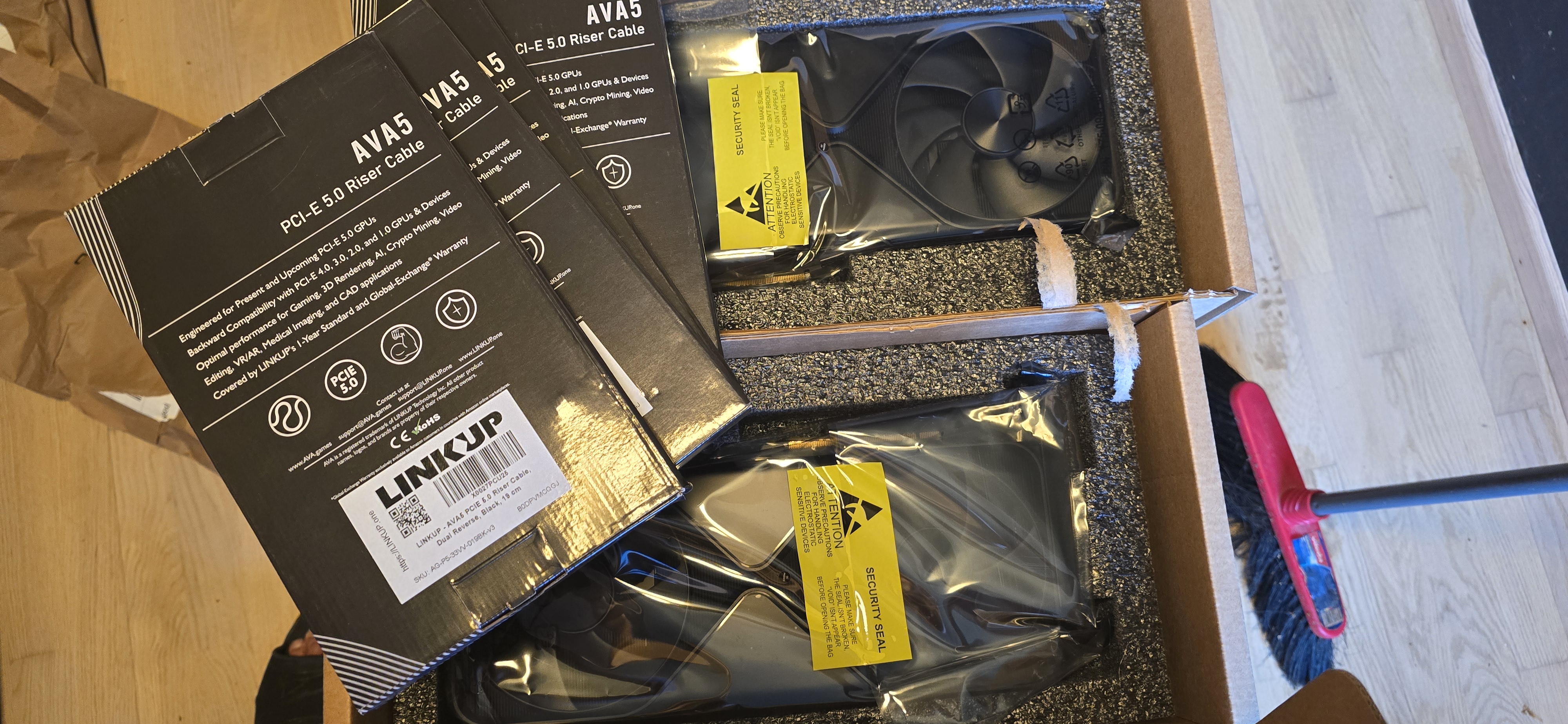LINKUP PCIe 5.0 riser cable boxes and two GPUs in anti-static bags with yellow security seals, on a wooden floor with a screwdriver.